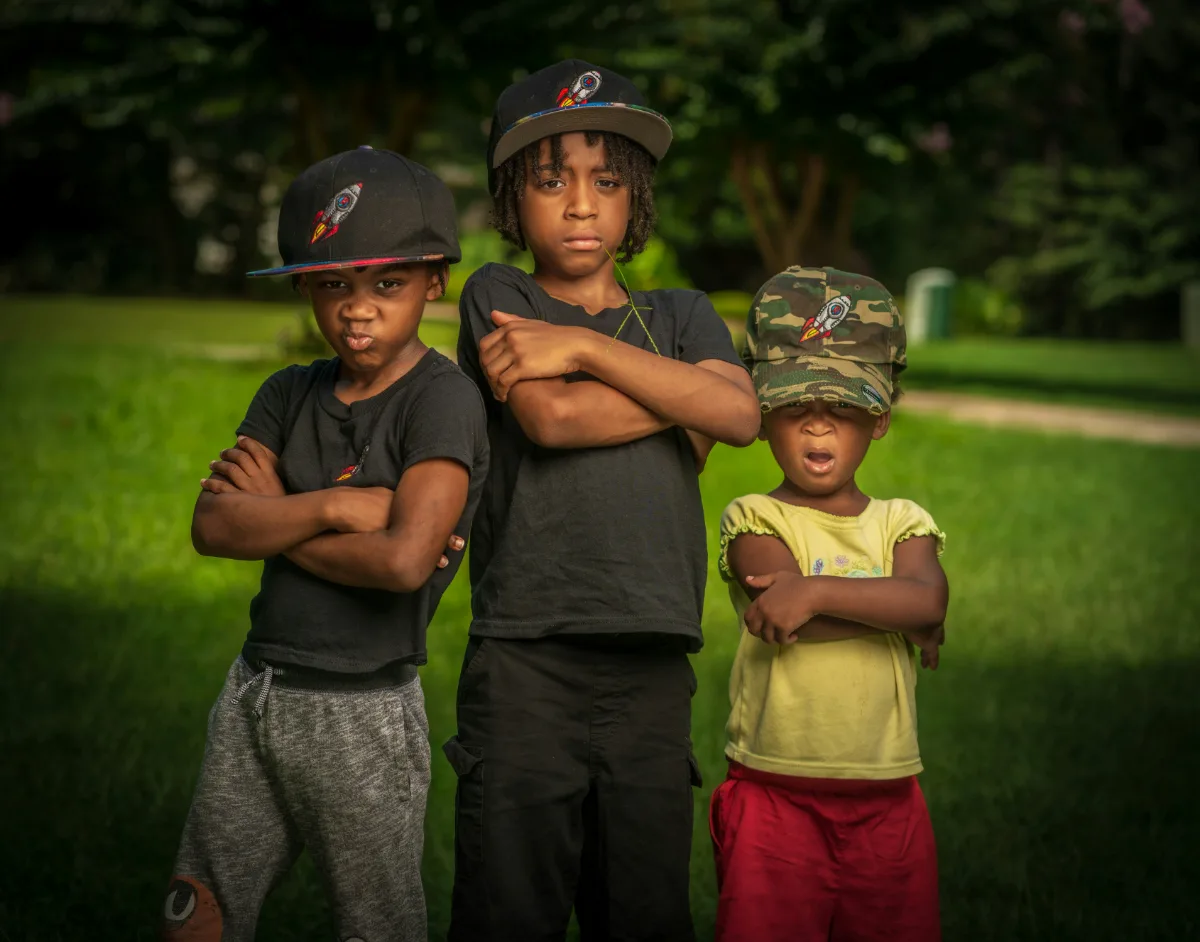 Three young children in baseball caps stand shoulder to shoulder on a lawn with their arms folded, doing their best serious faces.