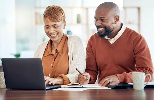 A couple smiling at a laptop at their kitchen table, one of them writing in a notebook.
