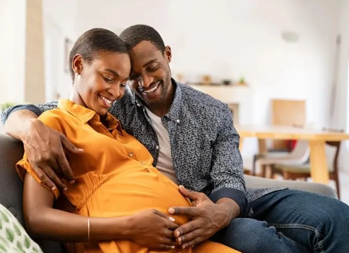 An expecting couple sit together on a sofa, his arm around her, both looking down at her hands resting on her bump.