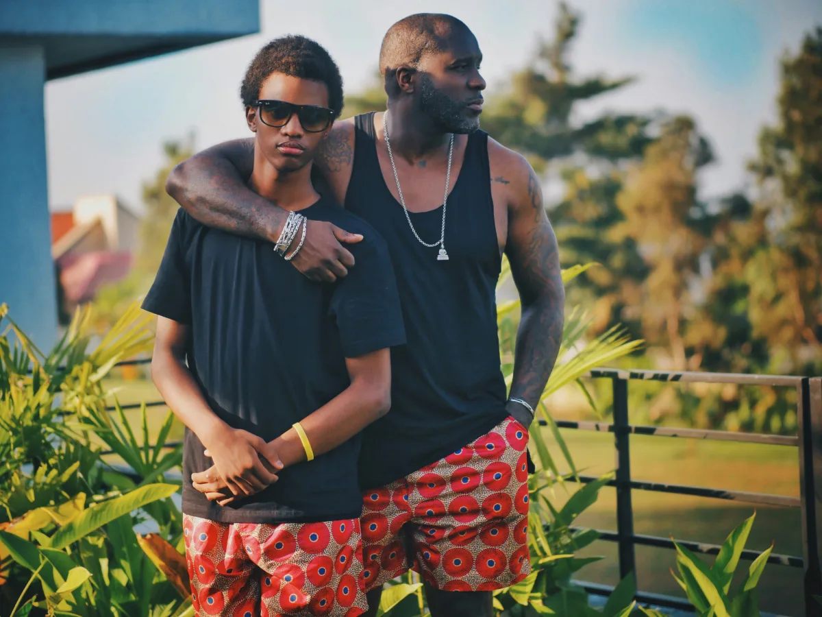 A father stands with his arm around his teenage son on a walkway, palms and a railing behind them.