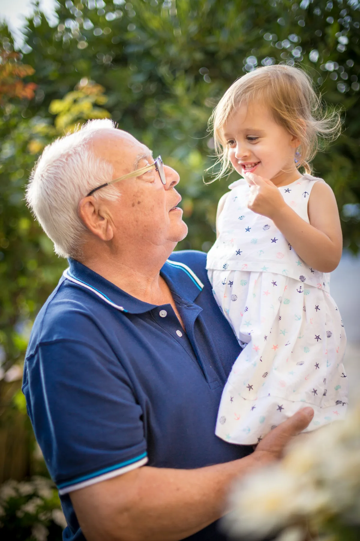 A grandfather in a navy polo shirt holds his small granddaughter at eye level in a garden, both of them mid-laugh.