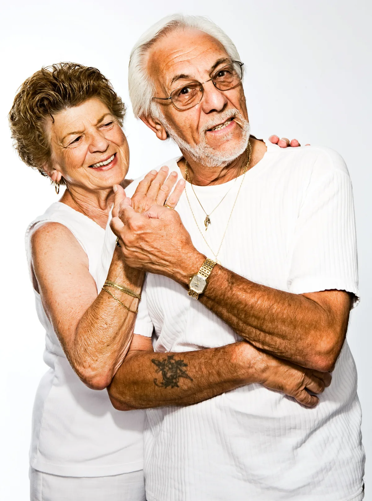 An older couple in white shirts stand close together against a plain background, her arms around him, both smiling at the camera.