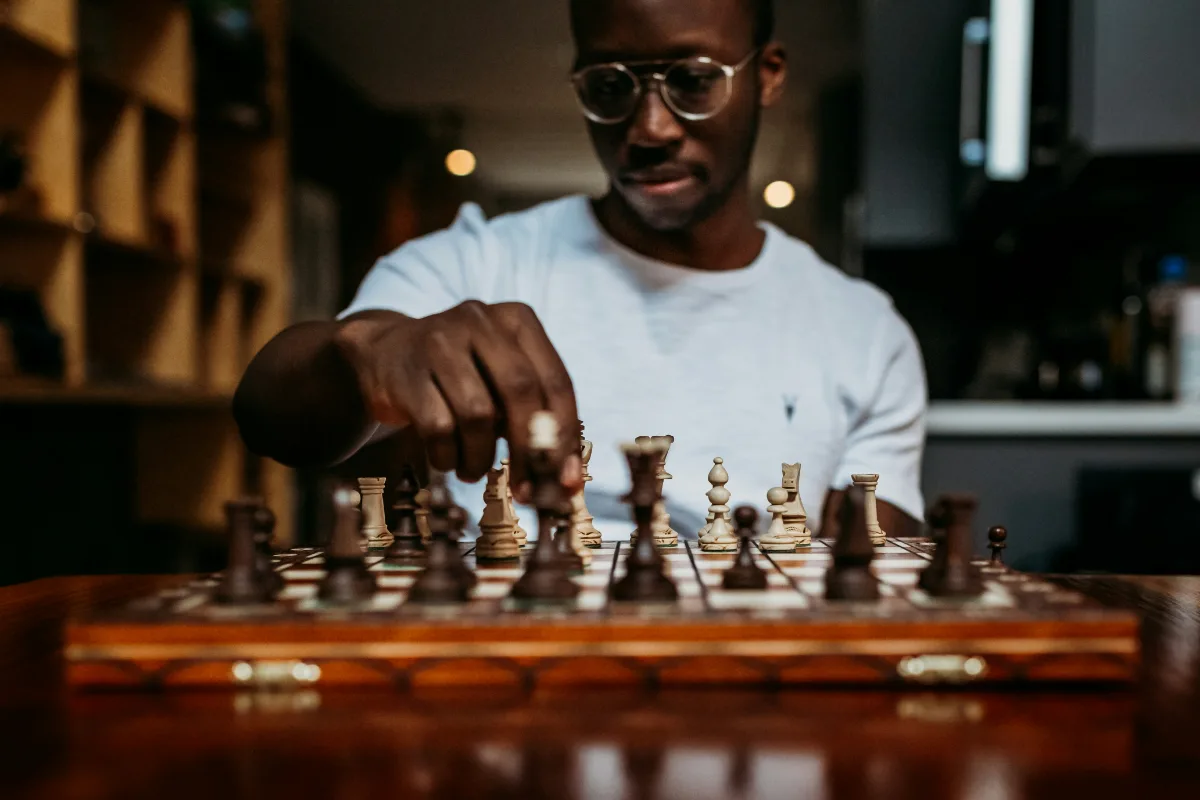 A young man in glasses leans over a wooden chess board, one piece held between his fingers mid-move.